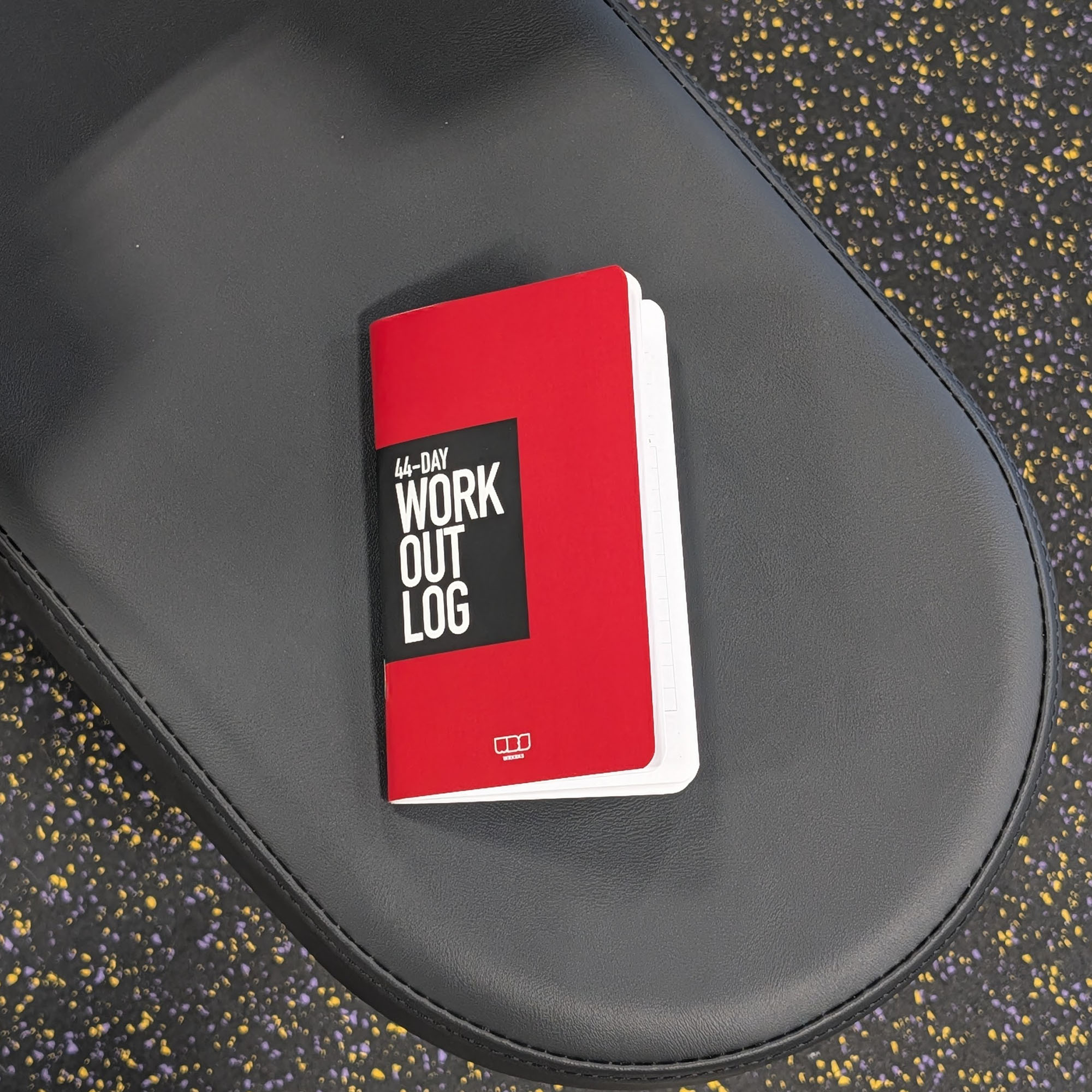 Red workout log book on a black gym bench with a speckled floor background