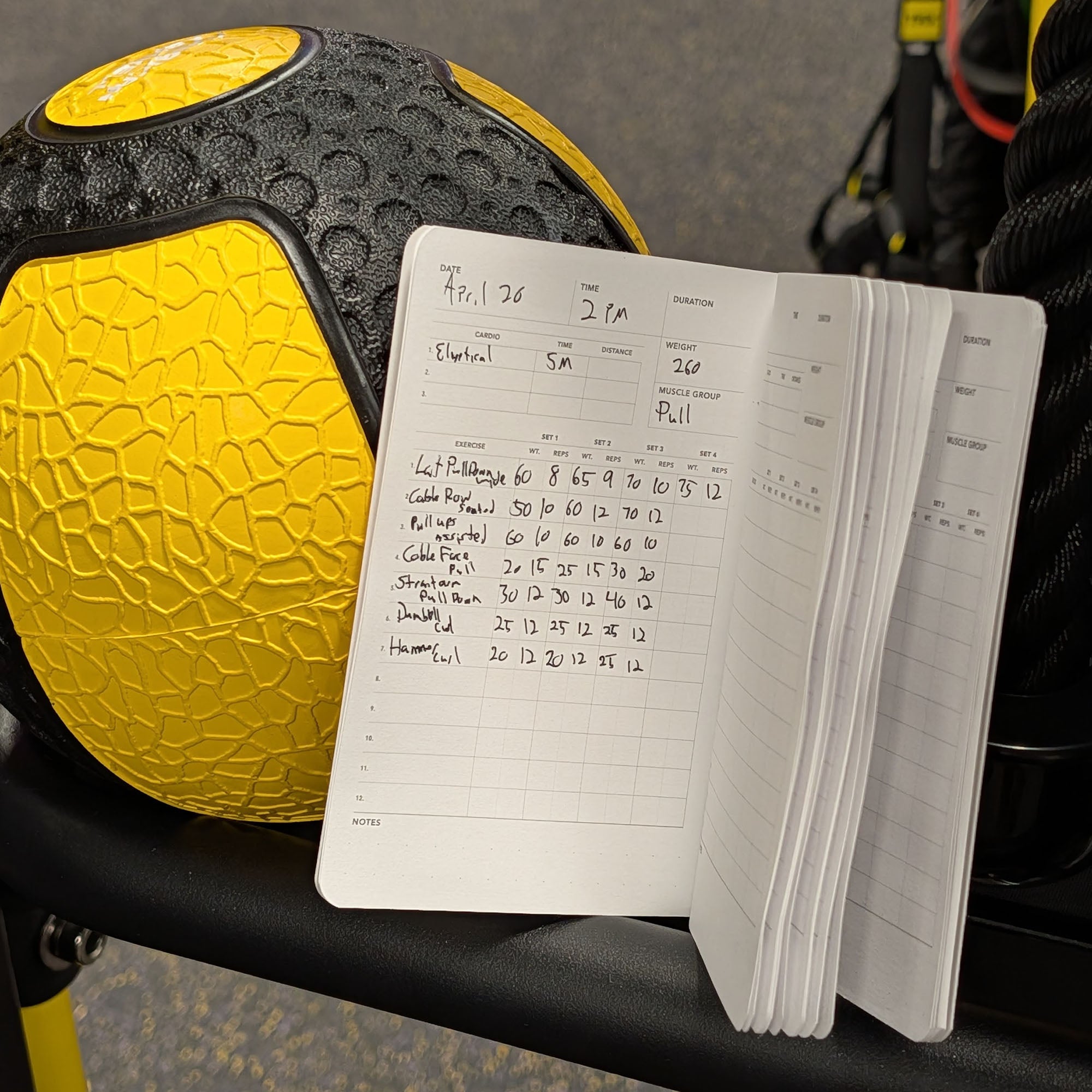 Open notebook with workout log next to a yellow and black medicine ball on a gym bench.